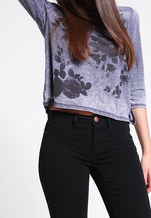 Lila, kurz geschnittenes Langarmoberteil mit Blumenmuster, kombiniert mit schwarzen Skinny Jeans. Das Oberteil hat eine lockere Passform und eine weiche Textur.