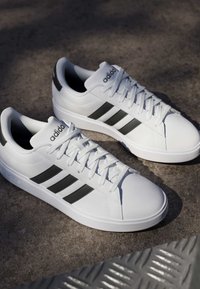 Weiße Adidas-Sneaker mit schwarzen Streifen und Schnürsenkeln, auf einer strukturierten Betonoberfläche im Sonnenlicht platziert.
