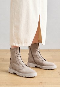 Beige nilkkurit, jotka on valmistettu sileästä nahasta, sisältävät nauhakiinnityksen edessä, pehmustetun kauluksen ja paksun valkoisen kumipohjan, jossa on sakara-kuvio.