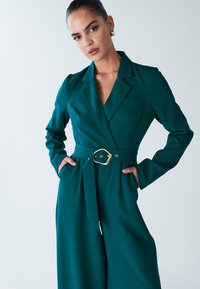 Teal jumpsuit with a fitted blazer top, long sleeves, and a wide waist belt featuring a gold accent, smooth fabric, and side pockets.