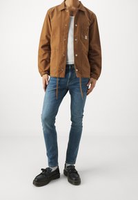 Man i en brun knäppt jacka över en vit skjorta, blå jeans, ljusgrå strumpor och svarta tofsloafers står framför en enkel vit bakgrund.