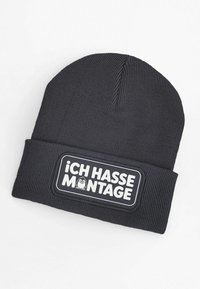 Black knit beanie with a ribbed texture and a patch featuring the text “¡ICH HASSE MONTAGE!” and a cartoon penguin.