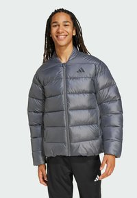 Uśmiechnięta młoda osoba z długimi dredami, ubrana w puszystą szarość kurtkę, stojąca na jasnym tle.