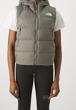 Personne portant un gilet matelassé gris sans manches avec capuche, zippé, par-dessus un t-shirt blanc à manches courtes et un legging noir avec logo sur la cuisse.