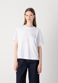 T-shirt en coton blanc à manches courtes, col rond, avec le logo "Calvin Klein" en teal et rose sur le côté gauche. Associé à un pantalon en denim noir.