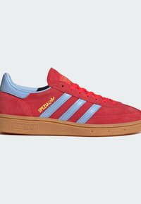 Červené semišové tenisky Adidas Spezial so svetlomodrými pruhmi, modrým opätkom, červenými šnúrkami, žltým logom a podrážkou z gumy na bielom pozadí.