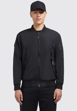 Schwarze Bomberjacke mit Reißverschlusstaschen, geripptem Kragen und Bündchen, aus glattem Nylonmaterial. Über einem schwarzen Outfit getragen.