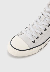 Grå canva sneakers med en hvit gummikappe, svart trim og hvite snører som går gjennom metalløye.