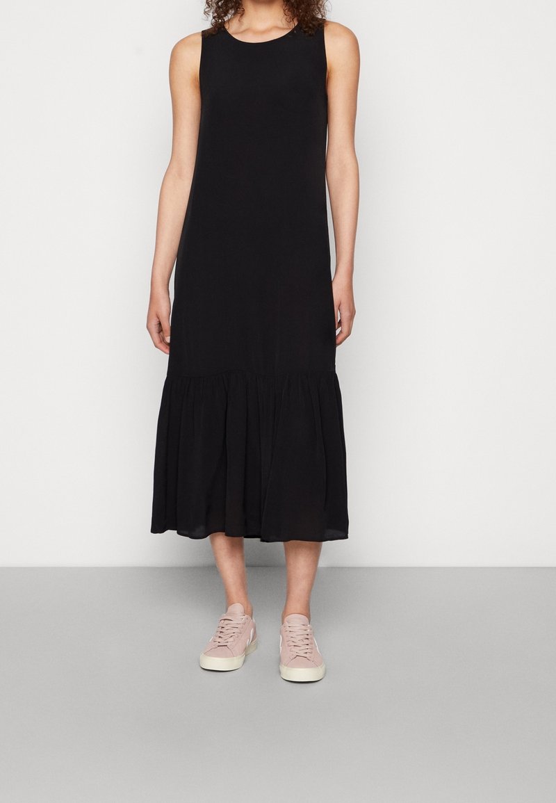 Femme portant une robe midi noire sans manches avec un ourlet volanté et des baskets roses, debout devant un fond clair uni.