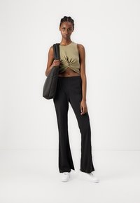 Olive green cropped tank top with a knotted front, paired with black flared pants and white sneakers. Model holds a dark tote bag.