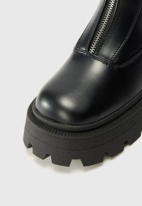Bota de cuero negro con suela gruesa de tracción y cremallera plateada frontal, mostrando un primer plano de la textura de la puntera y la suela sobre fondo blanco.