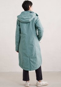 Lang lichtgroen winterjas met een capuchon, twee zijknopen en een afgeronde zoom. Draagt over donkere broek en witte sneakers.