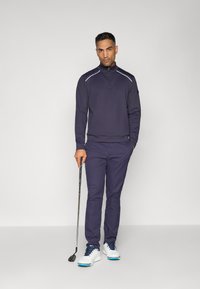 Tenue de golf marine comprenant un haut zippé avec des accents blancs, associé à un pantalon assorti et des chaussures blanches avec des détails bleus. Club de golf tenu à la main.
