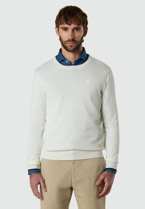 Mann mit braunen Haaren und Bart, der einen weißen Pullover mit Rundhalsausschnitt über einem blauen Hemd und beigen Hosen trägt, vor einem einfarbigen Hintergrund stehend.