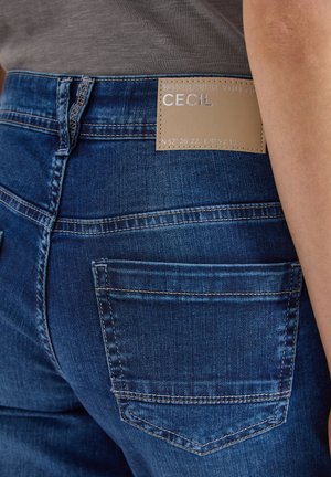 Close-up van de achterzak van een blauwe denim jeans met een beige label waarop staat "WHEREVER YOU GO CECIL" en geografische coördinaten.