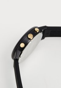 Montre analogique noire avec un cadran rond argenté. Équipée de boutons dorés et d'un bracelet noir texturé. Design minimaliste sans motifs visibles.
