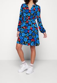 Blommig klänning med långa ärmar, prydd med blåa och röda blommor på en svart bakgrund, v-ringning och utsvängd kjol; matchad med vita sneakers.