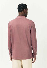 Vyras, atsukęs nugarą, vilki ilgomis rankovėmis melsvai violetinę marškinių ir šviesiai beige kelnes prieš paprastą baltą fono.