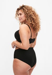 Set de lingerie noire comprenant un soutien-gorge à surface lisse et une culotte taille haute avec des accents en dentelle, vu de côté sur un fond blanc.