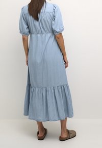 Kvinna klädd i en ljusblå midi-klänning med puffärmar och volangkanter, stående barfota mot en enkel vit bakgrund.