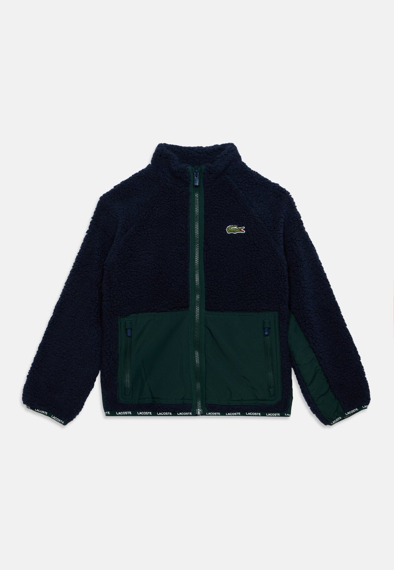 Μπλε μπουφάν fleece σε ναυτικό τόνο με πλήρες μπροστινό φερμουάρ, δύο τσέπες με φερμουάρ, πράσινο κάτω πάνελ και μικρό λογότυπο κροκοδείλου στο στήθος.