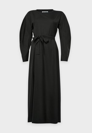 Modèle portant une robe noire à manches longues, nouée à la taille, et des bottines noires, sur un fond uni clair.