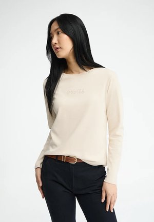 Frau mit langen schwarzen Haaren, die ein beigefarbenes Langarmshirt mit der Aufschrift "Polo Club" und schwarze Hose mit braunem Gürtel trägt, schaut nach links.