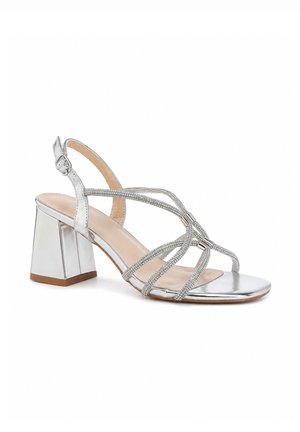 Sandale argentée avec talon bloc, multiples fines brides ornées de strass et une boucle de cheville réglable sur fond blanc.