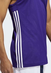 Homme portant un maillot de sport sans manches violet avec trois rayures blanches verticales sur le côté et un short blanc.