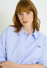 Camisa azul claro abotonada con mangas cortas, con un bolsillo en el pecho y texto bordado "Blue" en negro. Textura de tela suave.