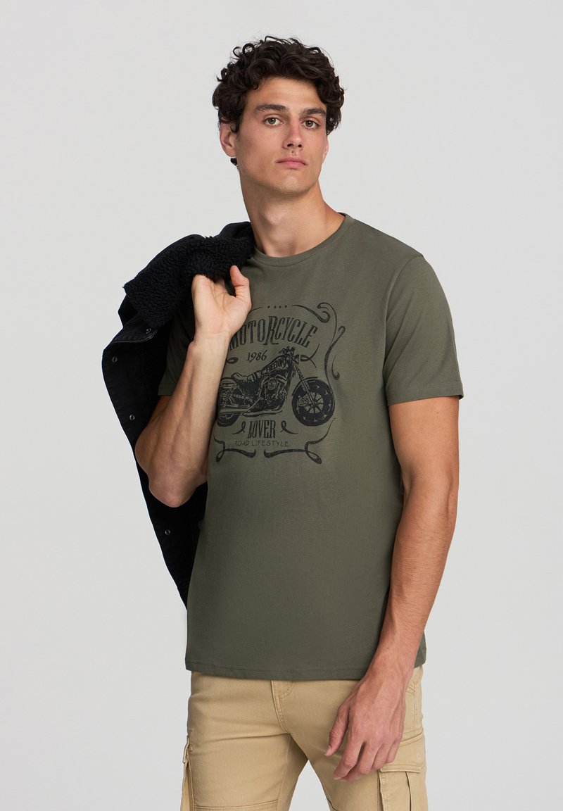 T-shirt in cotone verde oliva con una grafica di una motocicletta in nero. Il modello tiene un giubbotto foderato in sherpa nero su una spalla.