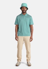 Tyrkysové polo tričko s límečkem, doplněné béžovými kalhotami. Model má na sobě odpovídající tyrkysový kbelíkový klobouk a světle šedé tenisky.