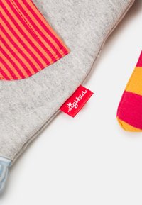 Grijze stof met een rood en oranje gestreepte patch; bevat een felrode label met witte tekst en een kleurrijke patroonaccent.