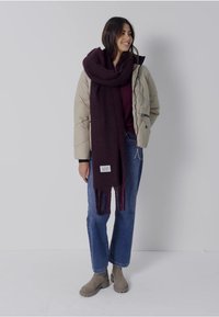 Sciarpa di lana bordeaux con bordi frangiati; indossata sopra un piumino beige e una maglia scura, con jeans blu e stivali ankle color cammello.