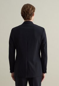 Giacca nera con un taglio sartoriale, apertura posteriore singola e maniche lunghe. Il materiale appare liscio con una texture minima, caratterizzato da polsini con bottoni.