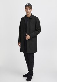 Mørk ullfrakk med krage-design, frontknapper og sidelommer, kombinert med en tettsittende svart skjorte, mørke bukser og en strikket lue.