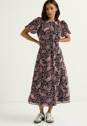 Donna con i capelli scuri che indossa un vestito midi nero con fantasia paisley rosa e maniche a sbuffo, abbinato a sneaker bianche e nere.