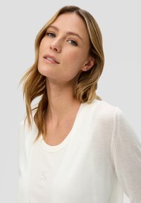 Witte lichte cardigan gelaagd over een crèmekleurig T-shirt, met een klein geborduurd logo. Zachte textuur met een ontspannen halslijn.