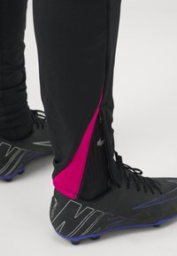Schwarze Sporthosen mit einem pinken Seitenpaneel, versehen mit einem Reißverschluss am Knöchel, kombiniert mit schwarzen Fußballschuhen mit blauen Akzenten.