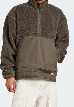 Person trägt einen dunkelolivgrünen Fleecepullover mit Viertelreißverschluss und weiße Hose, Hand in der Tasche auf hellem Hintergrund.