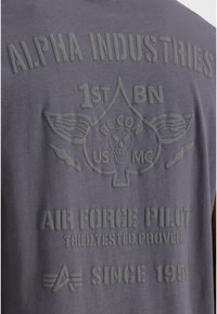 Camiseta gris con texto y gráficos en relieve, incluyendo "ALPHA INDUSTRIES", un emblema de calavera y motivos de aviación. Textura de tela suave.