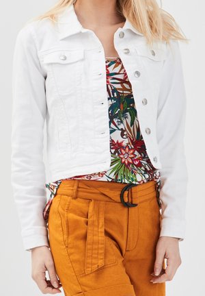 Femme portant une veste blanche à boutons, un haut à motifs floraux tropicaux, et un pantalon taille haute moutarde avec une boucle de ceinture noire.