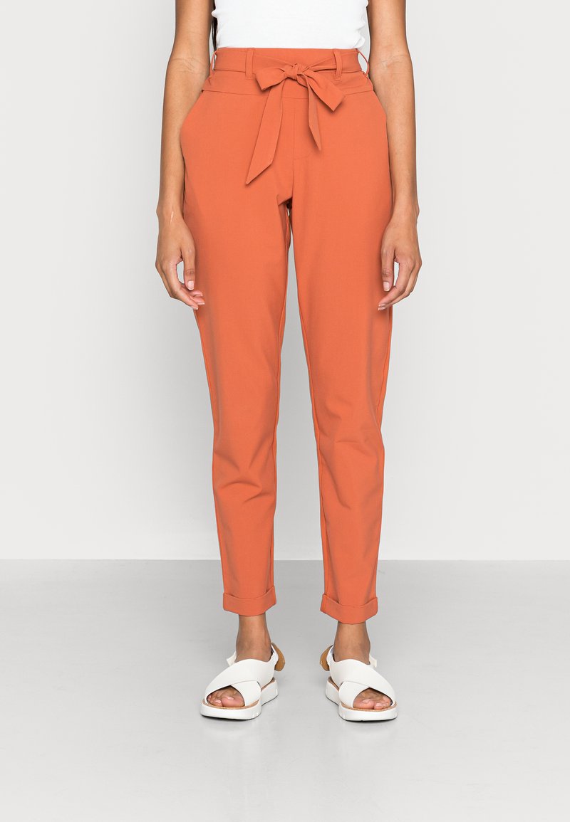 Calças ajustadas laranja com cinto de tecido, apresentando bainhas dobradas. Combinadas com sandálias brancas. Textura suave, design slim fit.