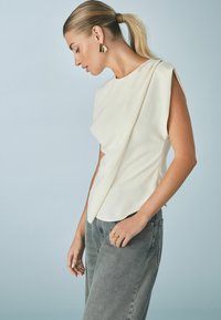 Blusa de crema con mangas en ángulo, textura suave y diseño estructurado, combinada con jeans de talle alto grises.