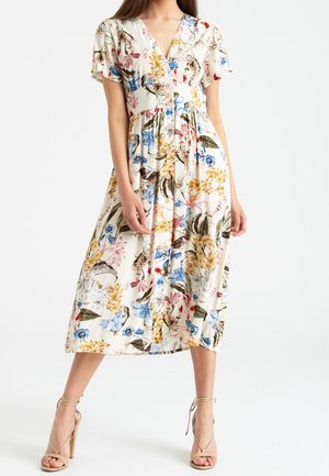 Femme portant une robe midi blanche avec un motif floral multicolore, manches courtes, boutons sur le devant, et des sandales à talons hauts beige.