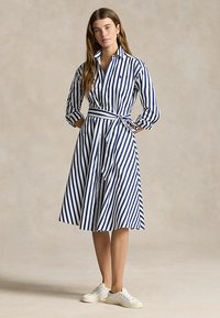 Jeune femme portant une robe chemise à rayures verticales bleues et blanches avec une ceinture à la taille et des baskets blanches, debout les mains dans les poches.