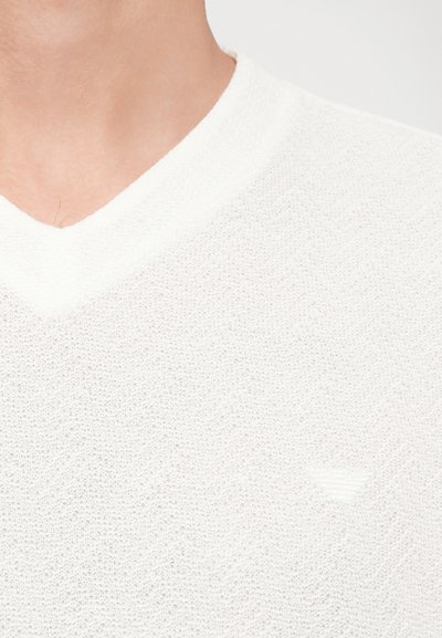 Valge V-kaelusega kampsun, millel on tekstuurne kootud muster koos peene chevroni disainiga ja väike logo rinnal.
