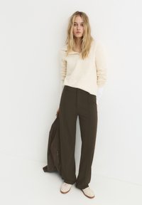 Maglione crema con colletto alto, pantaloni larghi verde oliva e sneaker bianchi. La modella tiene una giacca marrone, in piedi davanti a uno sfondo bianco.