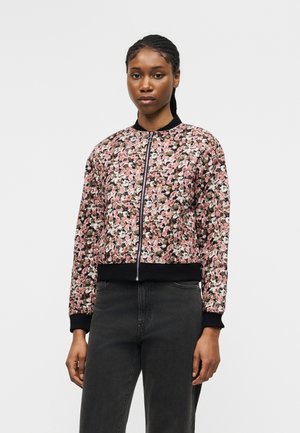 Kobieta w różowej, czarnej i białej bomberce w kwiaty na zamek błyskawiczny, z czarnymi mankietami i kołnierzem, w połączeniu z ciemnoszarymi dżinsami.