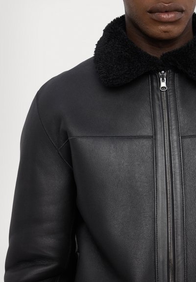 Veste en cuir noir avec un col en shearling texturé noir, fermeture éclair à l'avant et détails de design structuré sur les épaules.